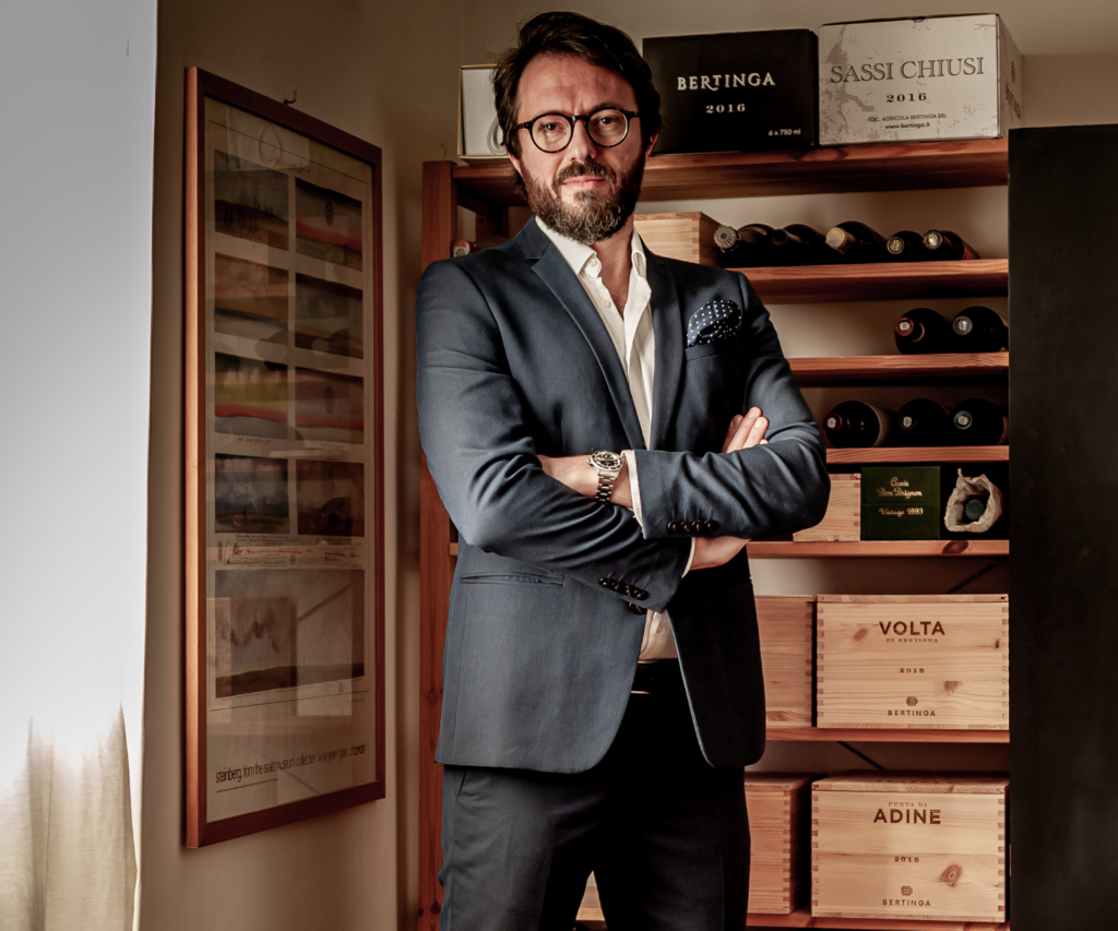 A man in a grey suit stands with his arms cross in a wine cellar. Behind him are cases of wine all adorned with the Bertinga logo.